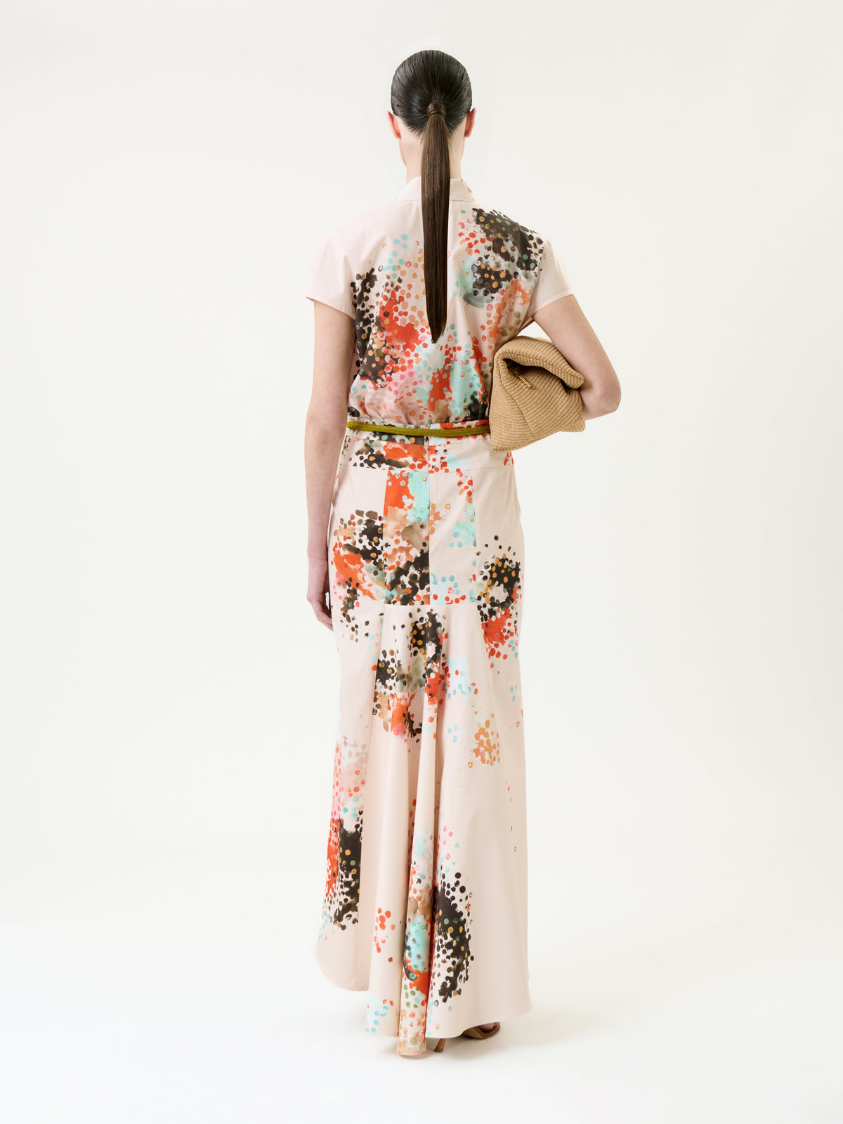 A woman with a ponytail, seen from behind, wears the Melia Skirt—a long pastel skirt with abstract patterns—and holds a woven clutch against a plain white background.