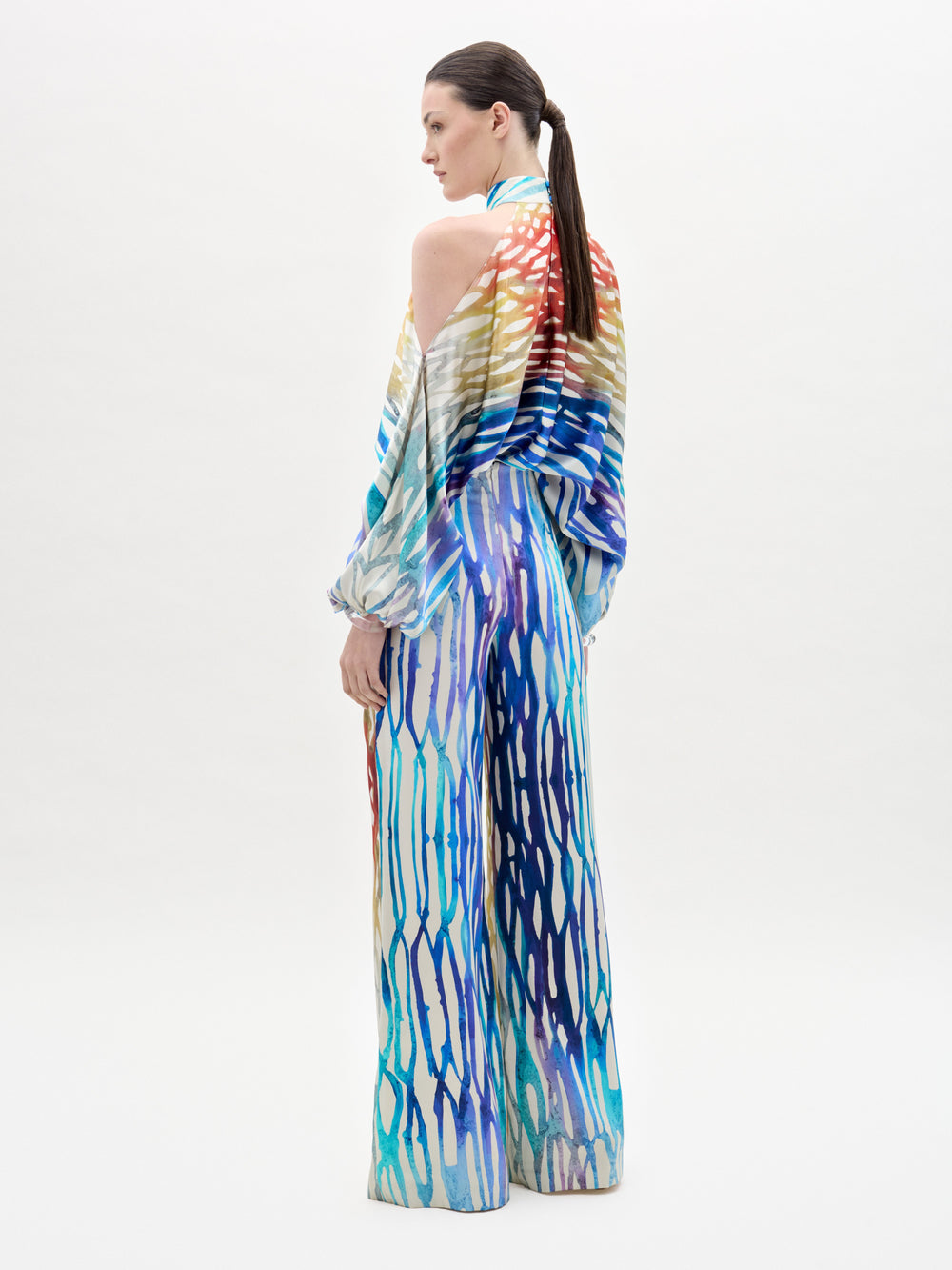 Woman poses against a plain background wearing the Andie Pant, a high-waisted, wide-leg style with colorful patterns, matching halter top, open shoulders, and long sleeves—all crafted from luxe Italian fabric.