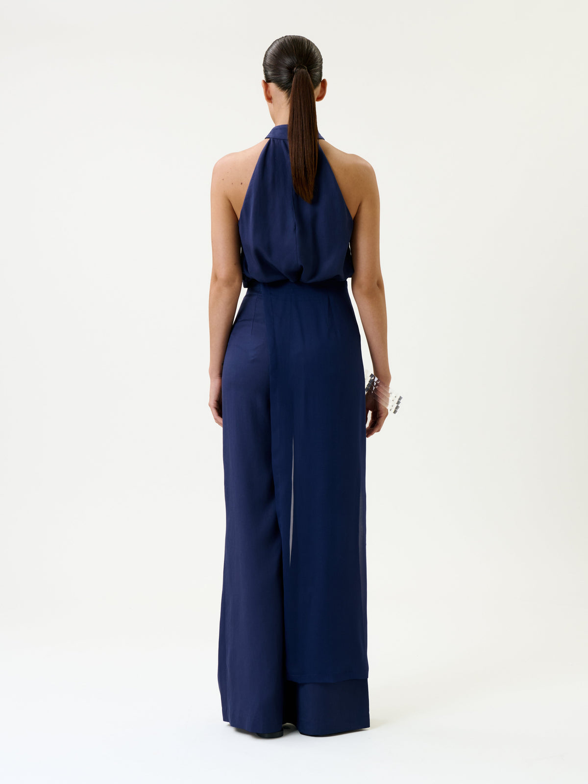 A person with long dark hair in a ponytail is standing with their back to the camera, wearing the Gaucin Pant in dark blue and a bracelet, against a plain light background.