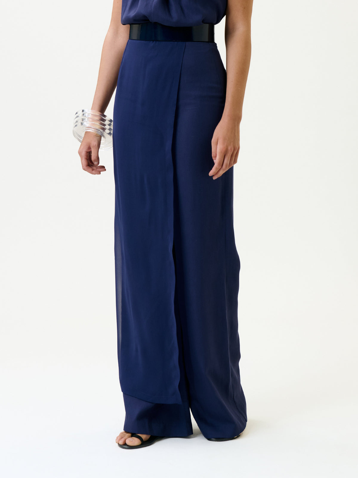 A person stands against a plain white background wearing the Gaucin Pant—navy blue, high-waisted, wide-leg trousers—paired with a matching top, black belt, clear chunky bracelet, and black open-toe sandals.