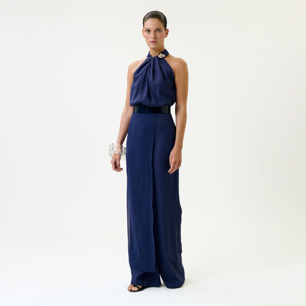 A woman stands against a plain background wearing the Gaucin Pant, styled with a sleeveless, navy blue halter top, wide black belt, black sandals, and silver bracelets.
