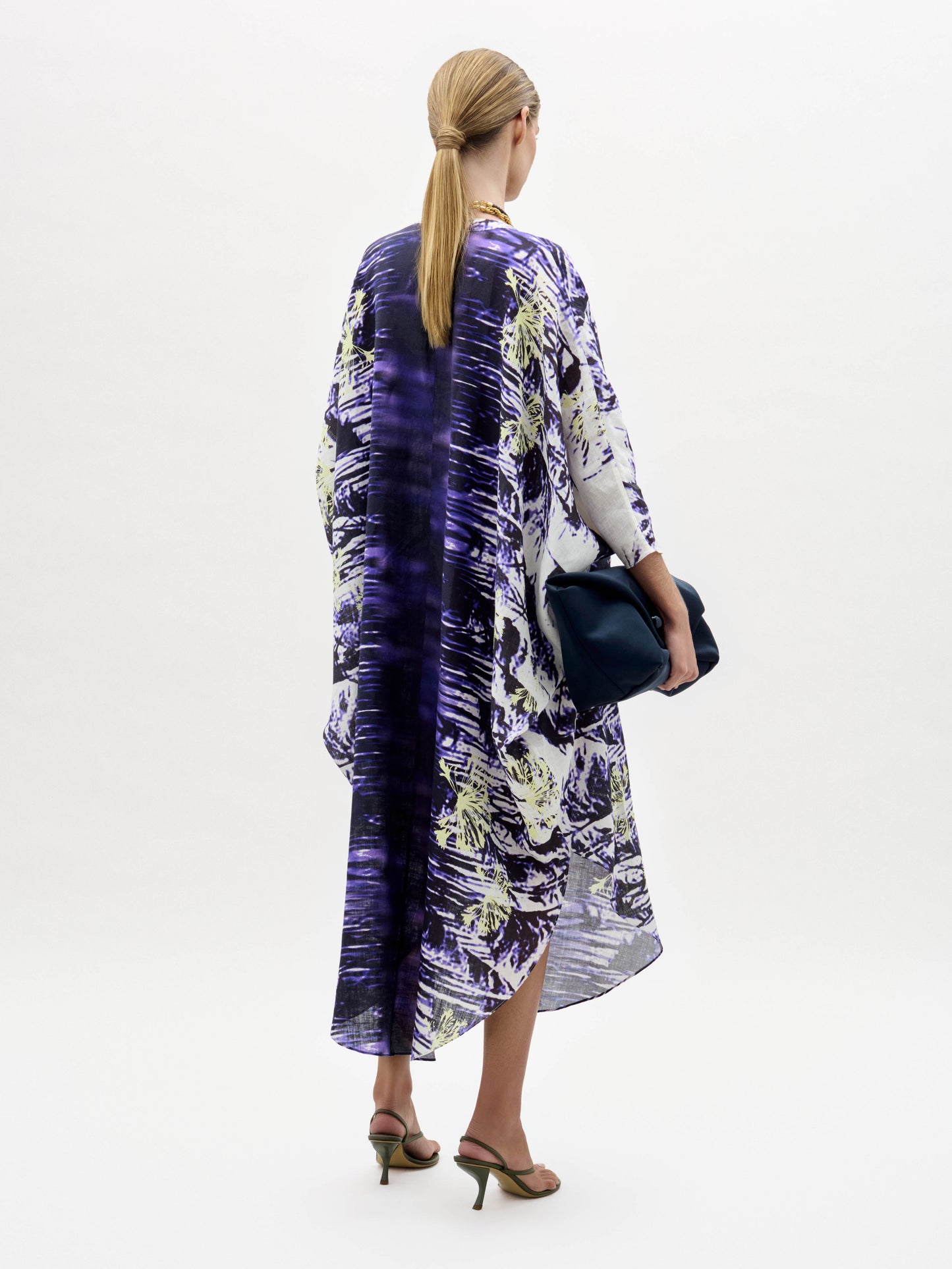 A woman stands against a plain white background wearing the Ilse Dress, a loose-fitting purple and white patterned piece, paired with a gold necklace, dark clutch, and heeled sandals.