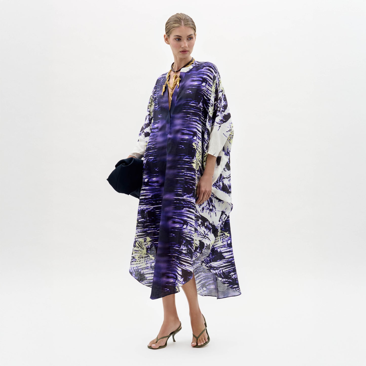 A woman stands against a plain white background wearing the Ilse Dress, a loose-fitting purple and white patterned piece, paired with a gold necklace, dark clutch, and heeled sandals.