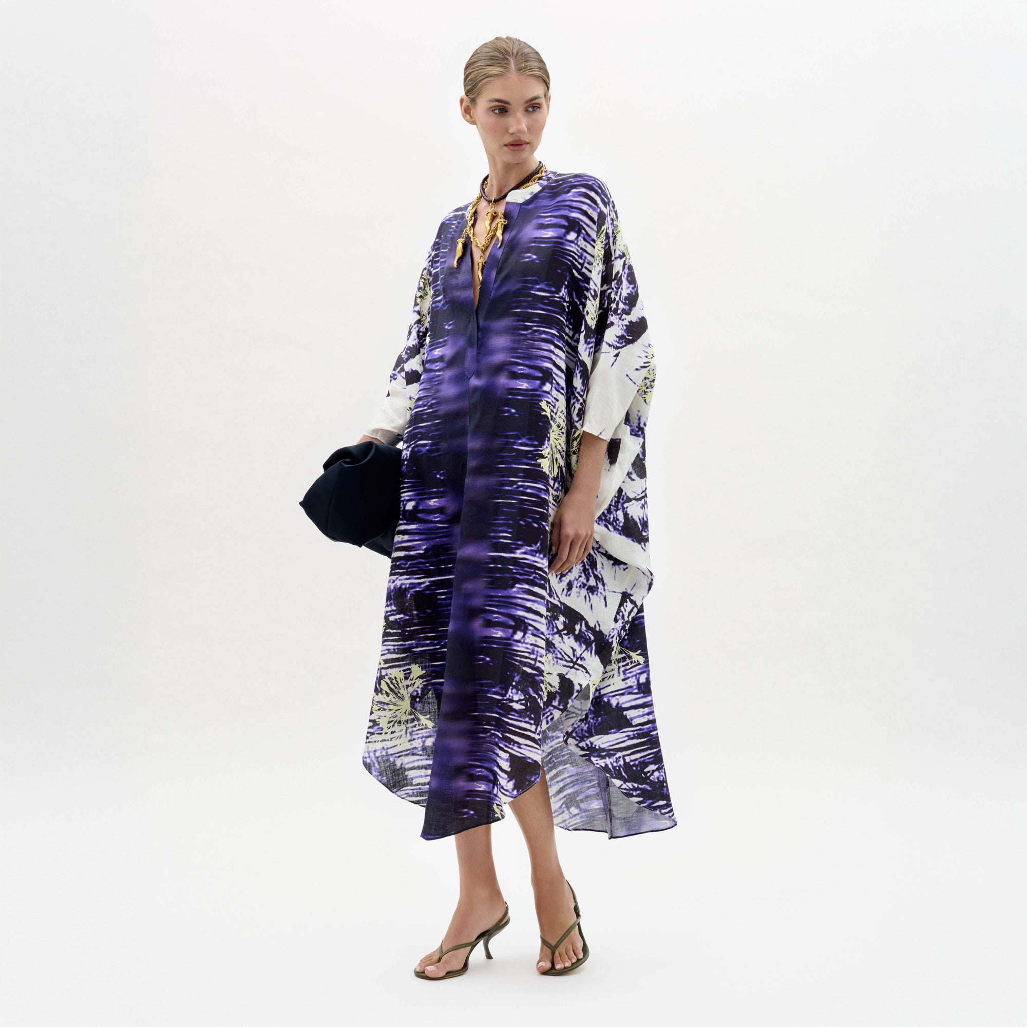 A woman stands against a plain white background wearing the Ilse Dress, a loose-fitting purple and white patterned piece, paired with a gold necklace, dark clutch, and heeled sandals.
