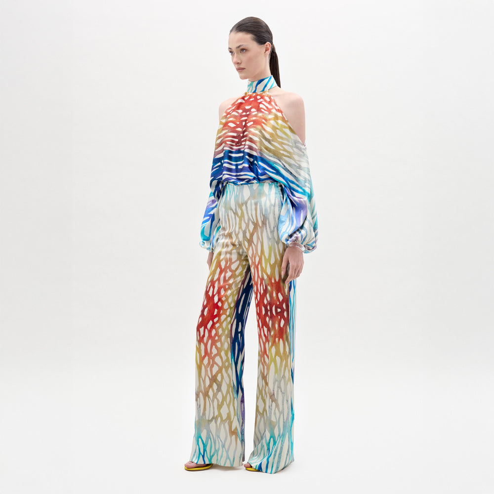 Woman poses against a plain background wearing the Andie Pant, a high-waisted, wide-leg style with colorful patterns, matching halter top, open shoulders, and long sleeves—all crafted from luxe Italian fabric.