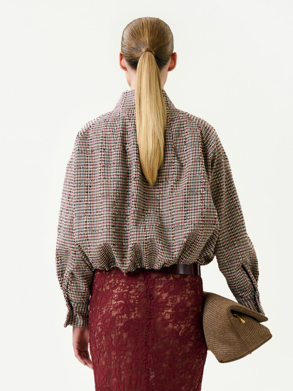A woman with a sleek ponytail is seen from behind, wearing the Julinka Jacket made in Colombia, tucked into a high-waisted burgundy lace skirt and holding a textured brown clutch.