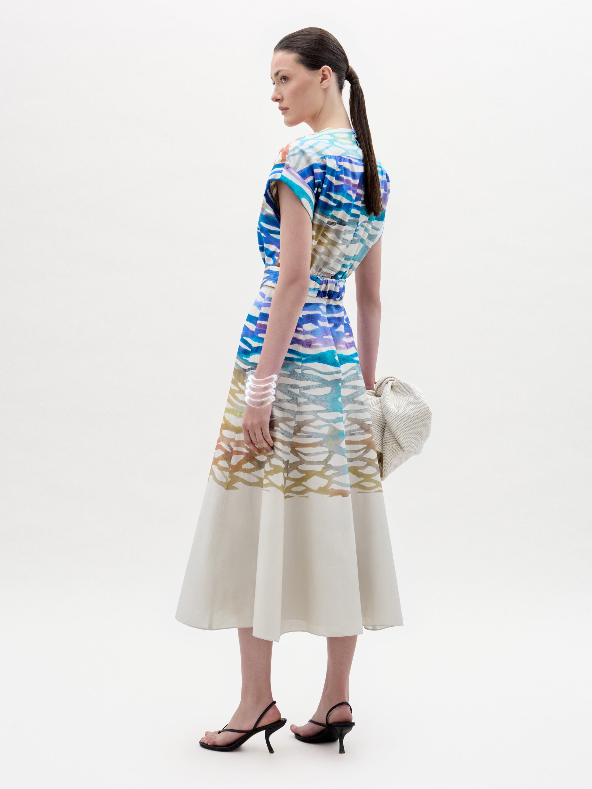 A woman stands against a white background wearing the Sakura Dress, a colorful patterned midi made from Italian fabric, paired with black heeled sandals and holding a large off-white bag.