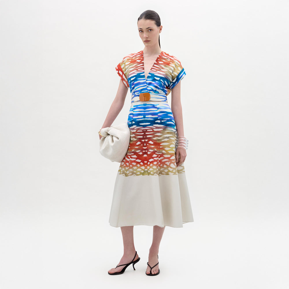 A woman stands against a white background wearing the Sakura Dress, a colorful patterned midi made from Italian fabric, paired with black heeled sandals and holding a large off-white bag.