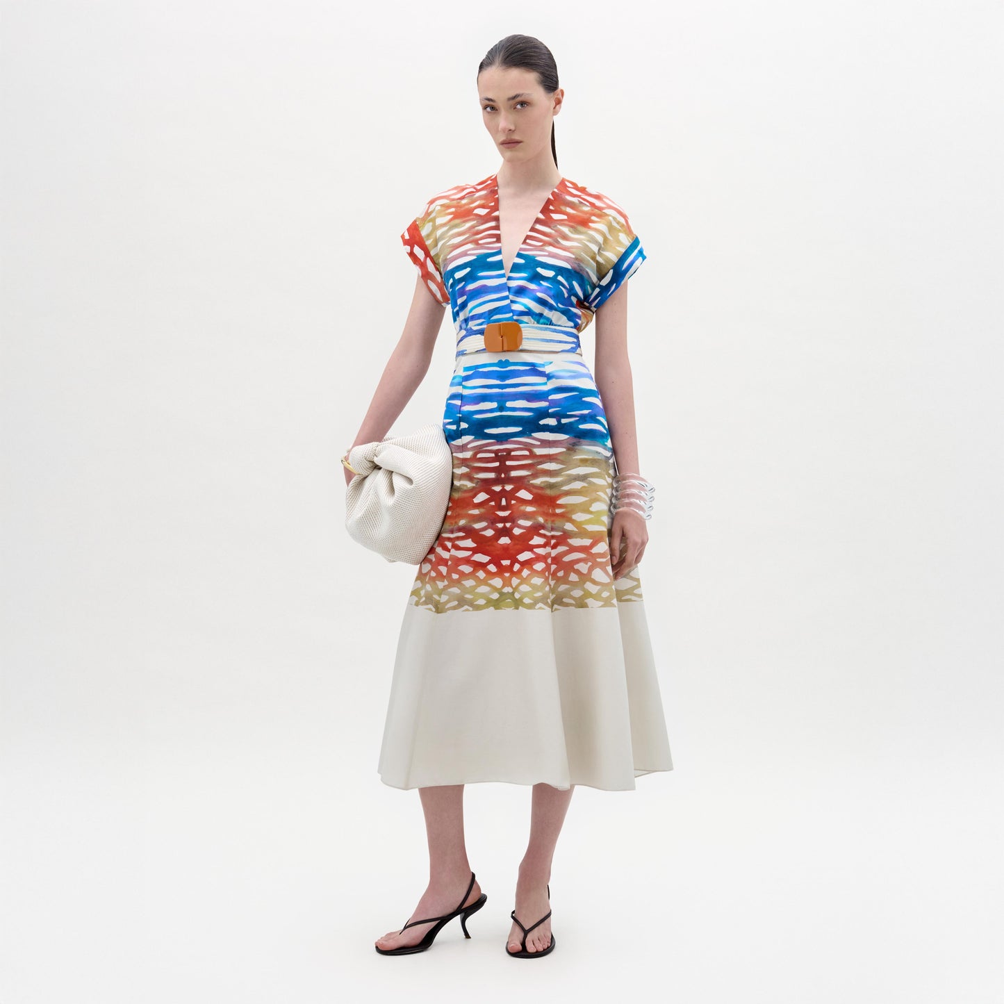 A woman stands against a white background wearing the Sakura Dress, a colorful patterned midi made from Italian fabric, paired with black heeled sandals and holding a large off-white bag.