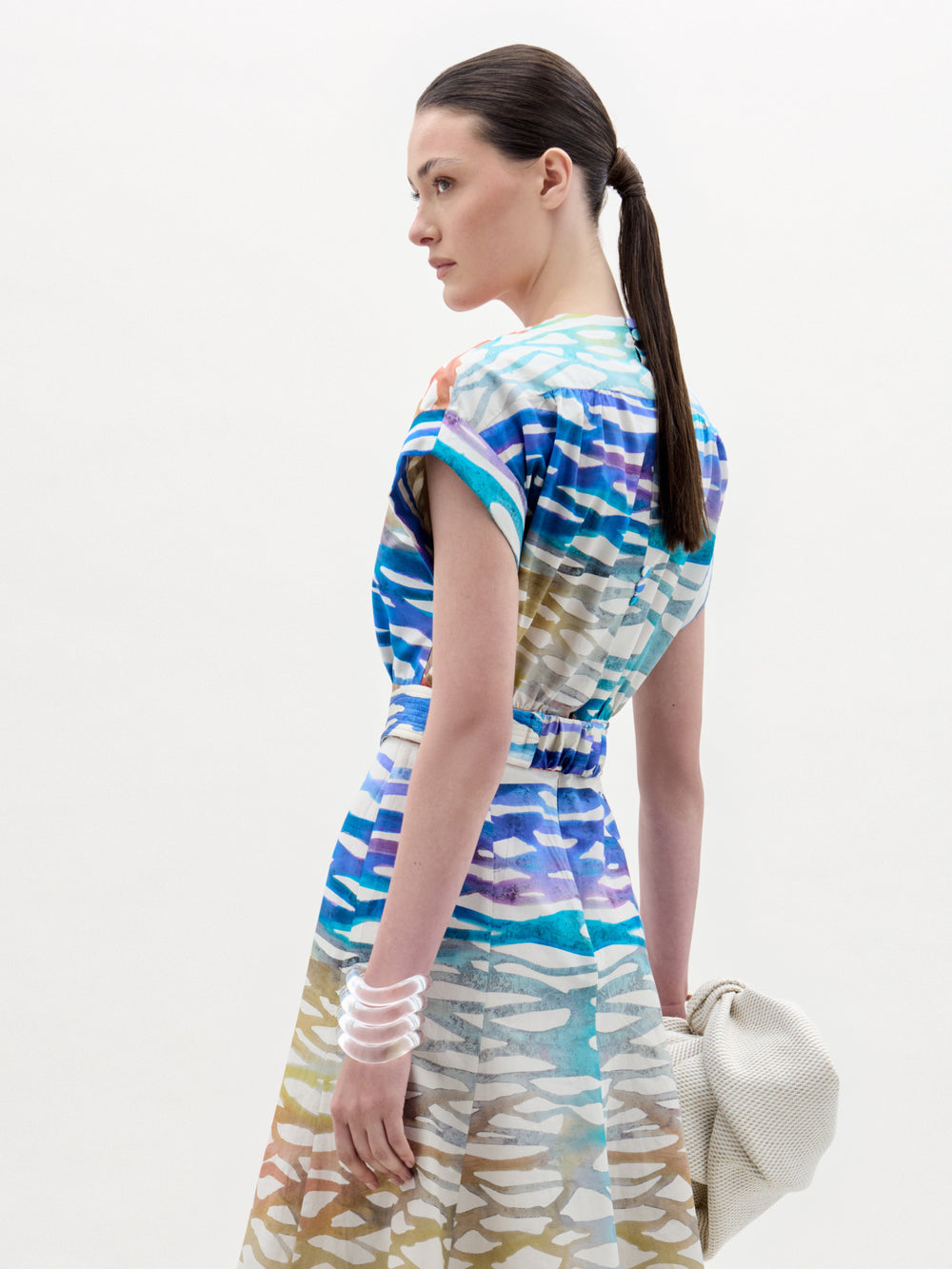 A woman stands against a white background wearing the Bara Blouse—crafted from 100% cotton Italian fabric—paired with a matching patterned beige skirt, wide spiral-buckle belt, and holding a textured cream bag.