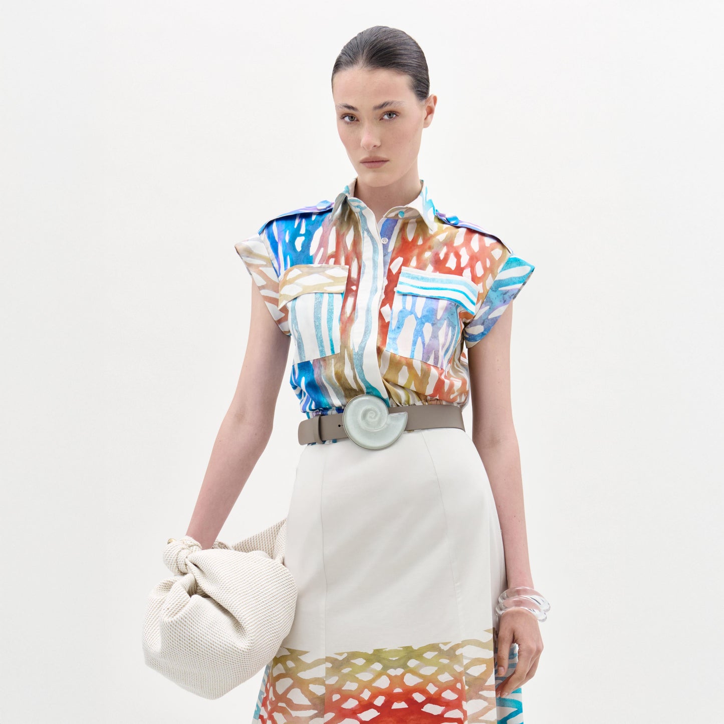 A woman stands against a white background wearing the Bara Blouse—crafted from 100% cotton Italian fabric—paired with a matching patterned beige skirt, wide spiral-buckle belt, and holding a textured cream bag.