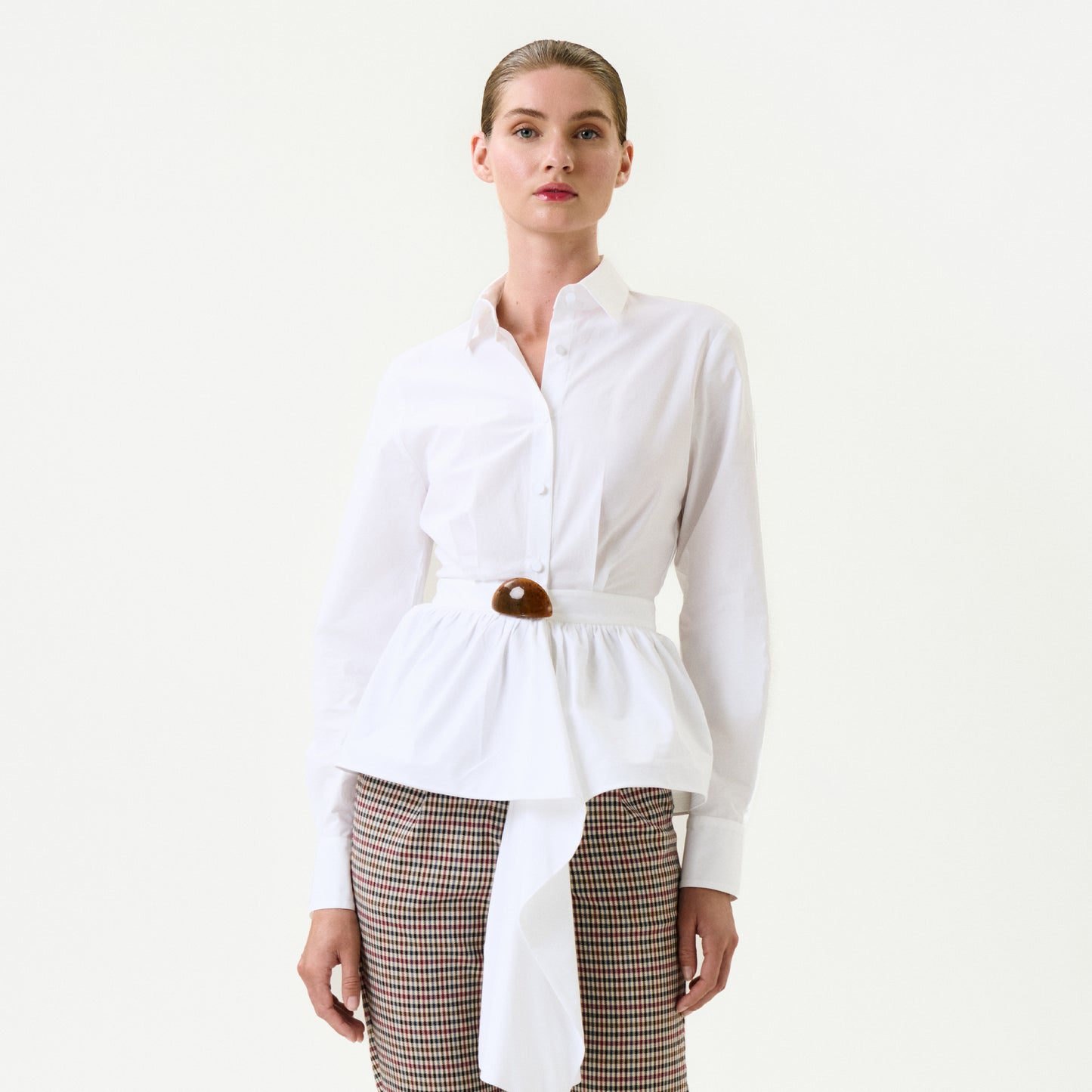 A woman with slicked-back hair and a neutral expression wears the Malina Shirt—a white 100% cotton button-down with peplum and brown belt—paired with high-waisted checked trousers. Photographed against a plain white background. Made in Colombia.
