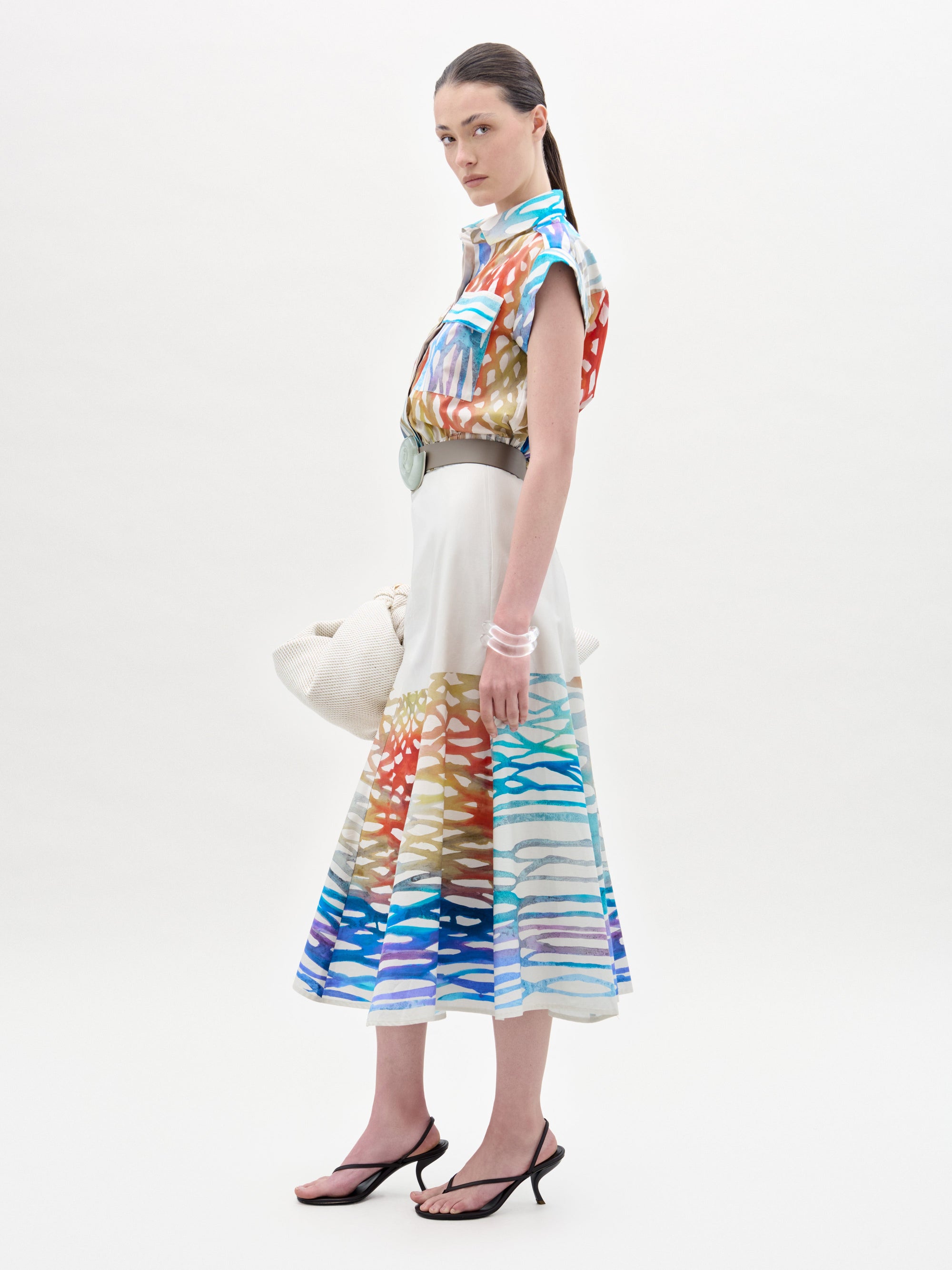 A woman wears the Erin Skirt, a colorful, abstract-patterned midi with an A-line silhouette, paired with a matching blouse, wide belt, white bag, clear bangles, and black sandals against a plain background.