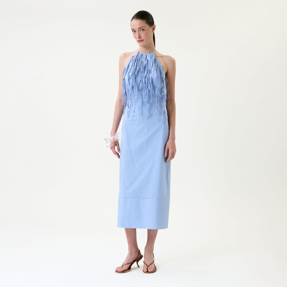 A woman stands against a plain background wearing the Otilia Dress, a light blue sleeveless midi dress featuring fringe detailing on the front, paired with brown heeled sandals. Her hair is tied back and she accessorizes with a white bracelet.