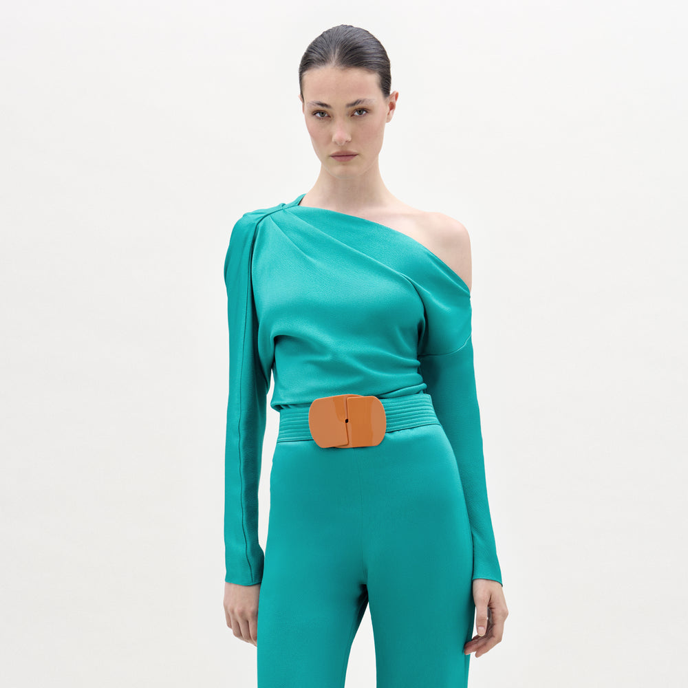 A woman wears the Viola Blouse in teal, an asymmetric design with draped details, paired with matching high-waisted pants and an oversized orange belt, all crafted from premium Italian fabric against a plain light backdrop.