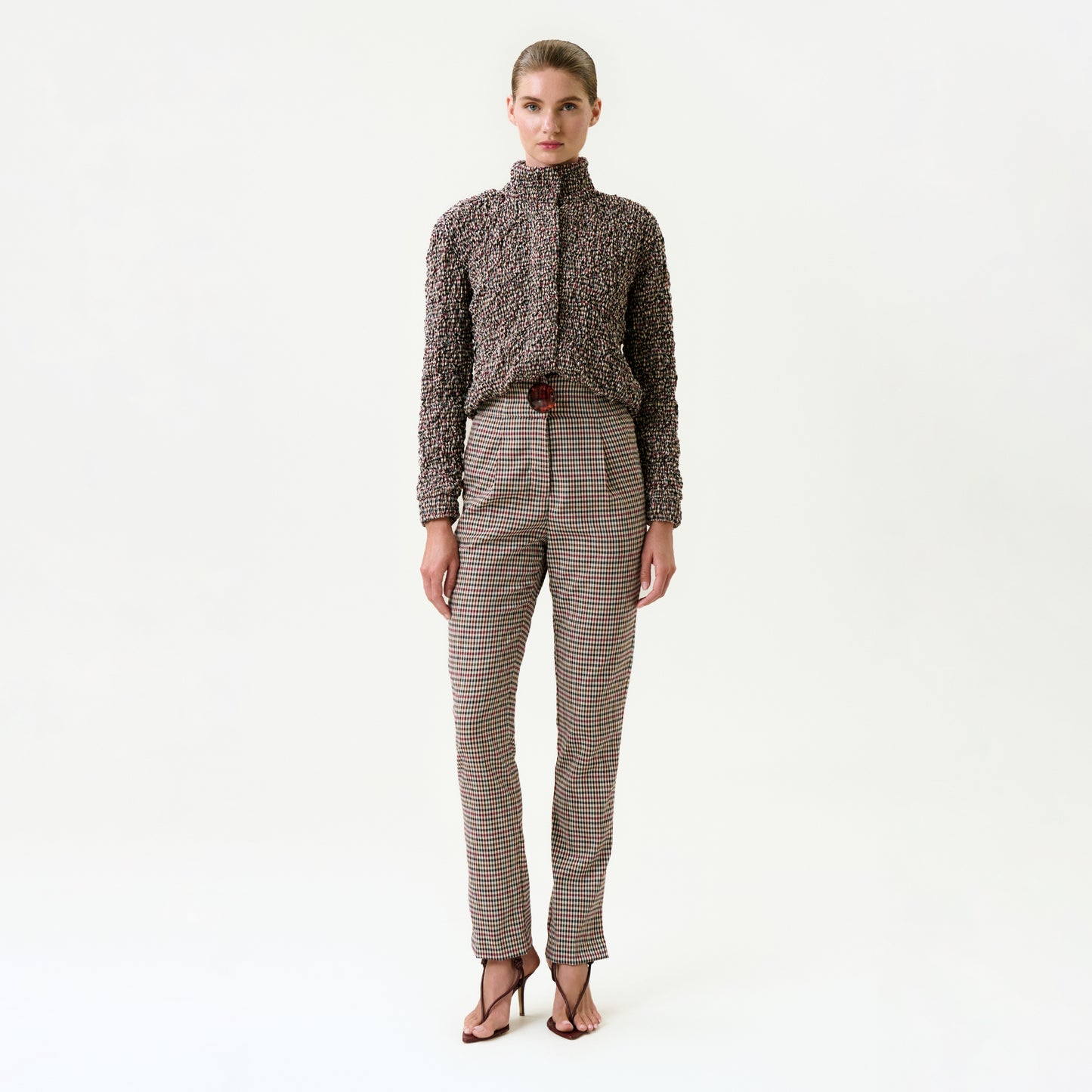 A woman stands against a white background wearing the Luca Pant—high-waisted, belted plaid pants made from Italian fabric for a true-to-size fit—with a textured long-sleeve top and open-toed heels; her hair is pulled back.