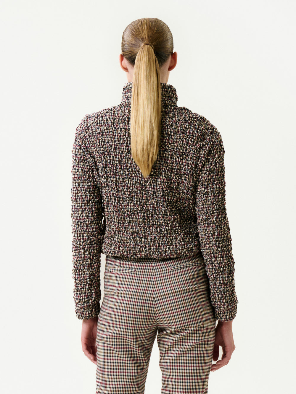 A person with long, straight blonde hair in a ponytail is shown from the back wearing the Zalta Jacket over a textured high-neck sweater and plaid pants, standing against a plain white background.