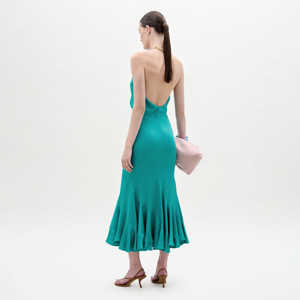 A woman with long dark hair in a low ponytail wears the Marlene Skirt in teal, paired with brown heeled sandals and a light pink clutch. She stands against a plain, light background, facing away.