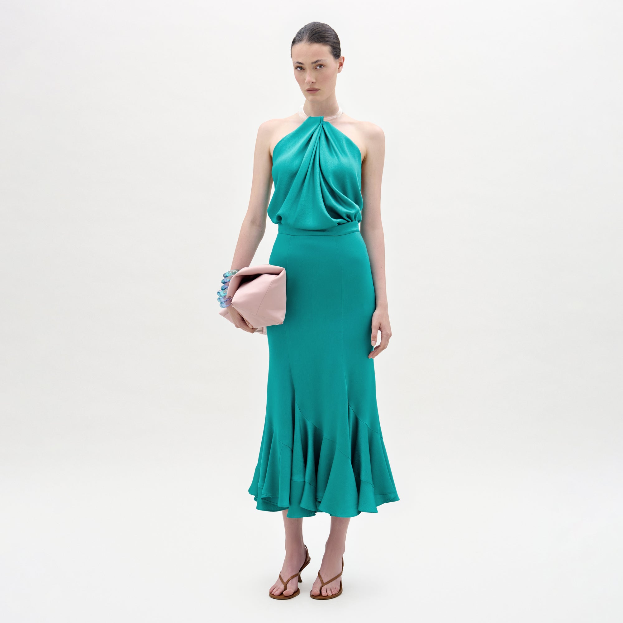 A woman stands against a plain white background wearing the Marlene Skirt, a teal, draped midi skirt with an A-line silhouette and ruffled hem, paired with brown heeled sandals and holding a large pale pink clutch.