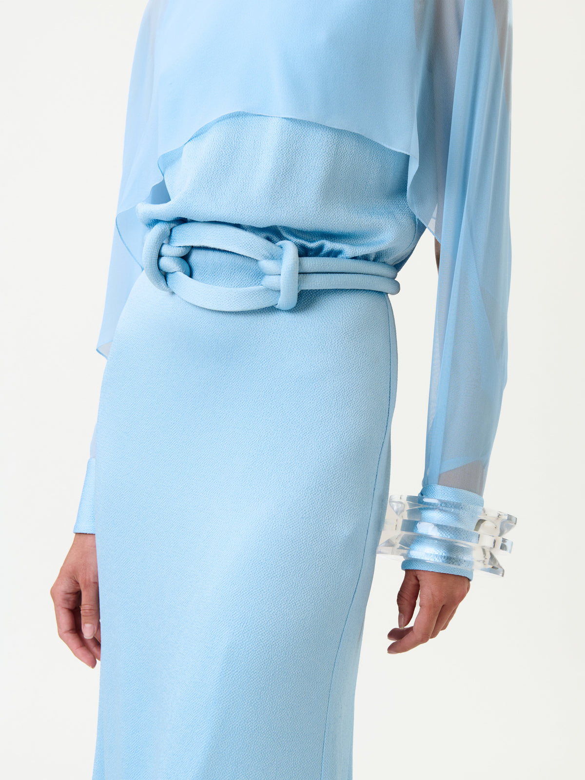 Model wearing the Rubina Dress in light blue, featuring a textured fabric, matching fabric belt, and sheer long sleeves, styled with clear chunky bracelets against a plain white background.