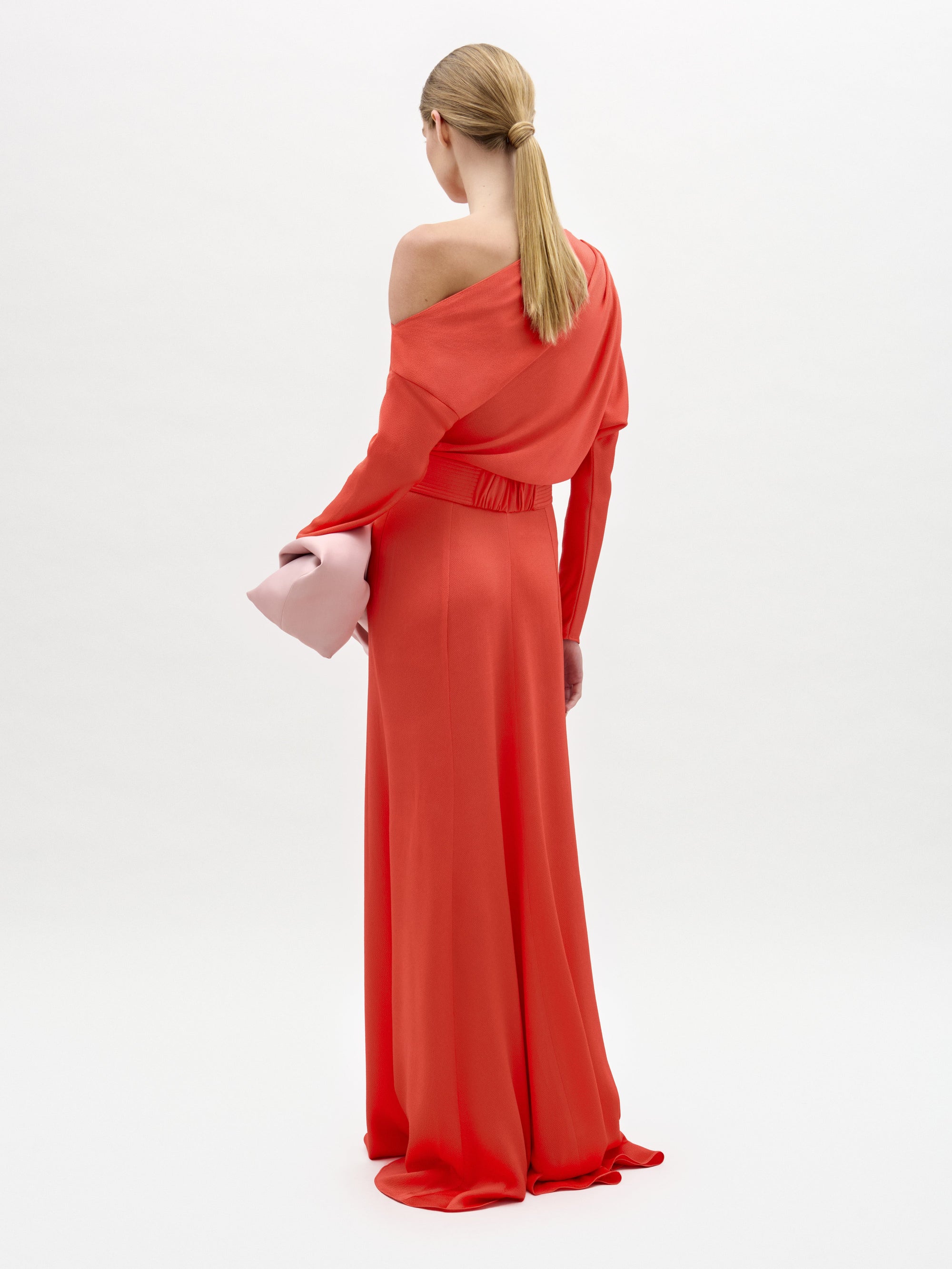 A woman stands against a white background wearing the Noelia Dress, a long, bright red maxi with an asymmetrical column silhouette, and holds a large pale pink clutch.