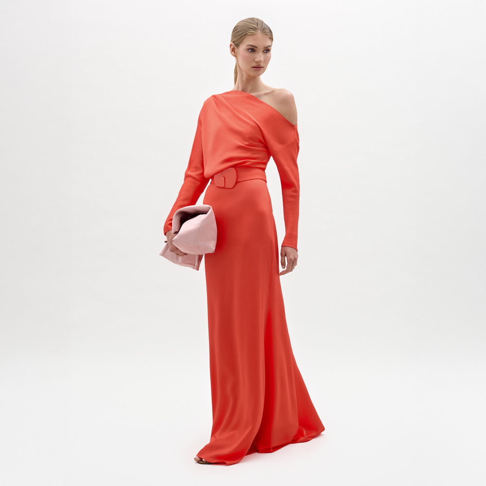 A woman stands against a white background wearing the Noelia Dress, a long, bright red maxi with an asymmetrical column silhouette, and holds a large pale pink clutch.