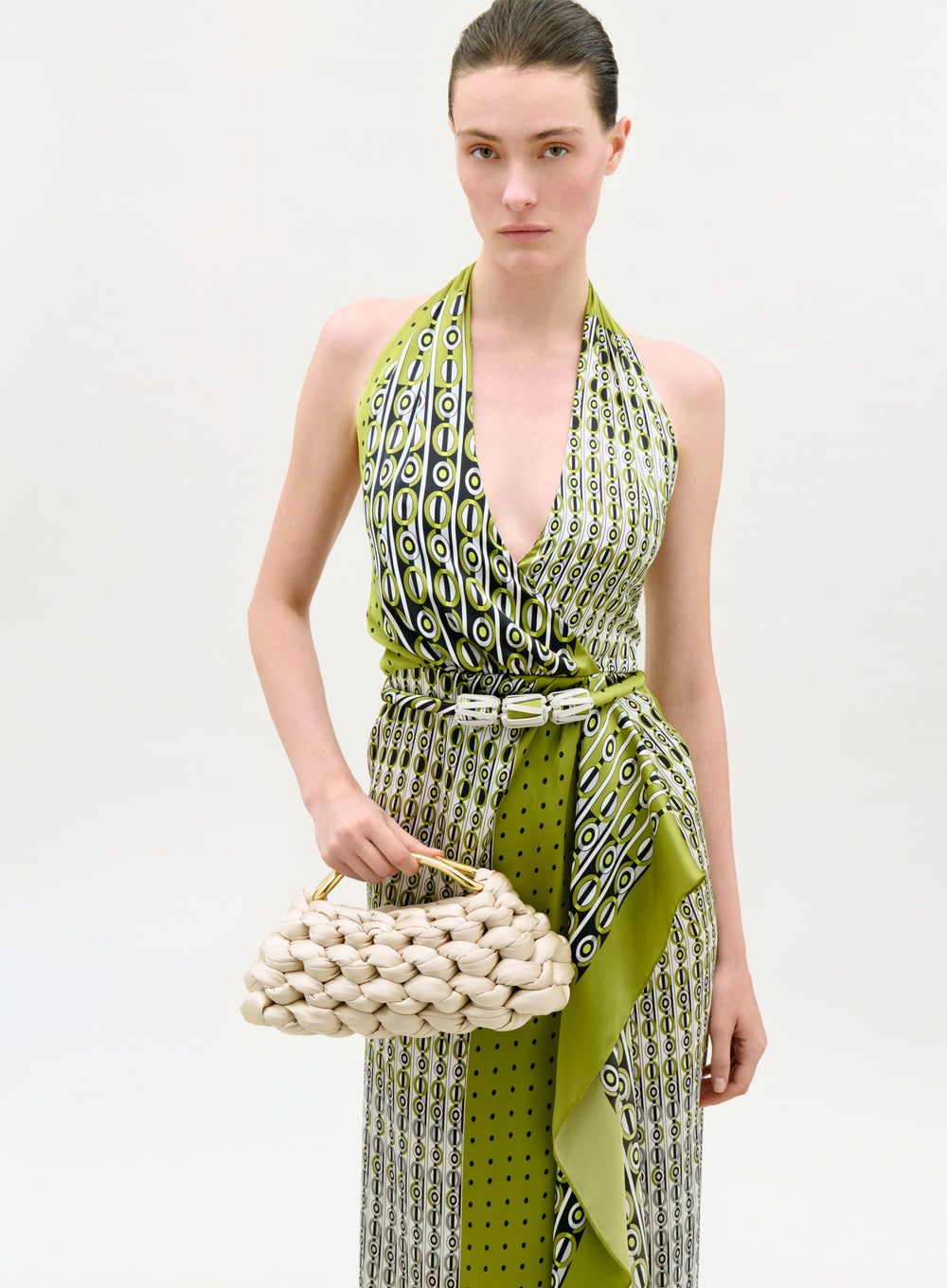 Woman wearing a green and white patterned dress holding a woven handbag on a white background