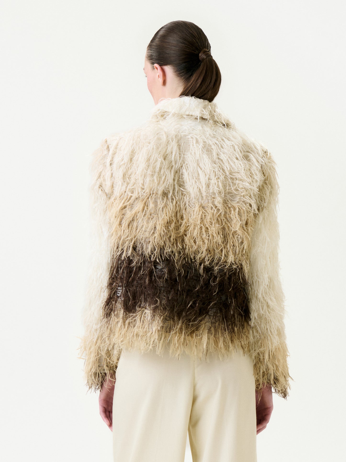 A person with dark hair in a low ponytail is shown from the back, wearing the Columba Jacket—an Italian fabric piece in shaggy cream and brown tones—paired with true-to-size cream pants, set against a plain light background.