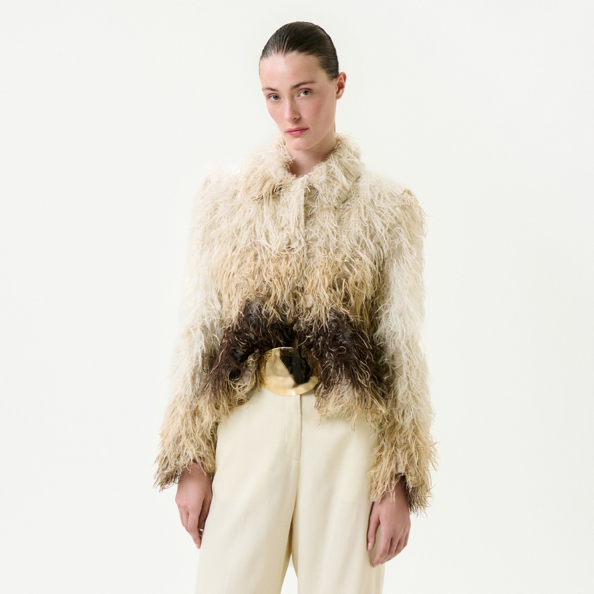 A person with slicked-back hair wears the Columba Jacket, a textured, shaggy cream and brown fur Italian fabric piece, paired with high-waisted cream pants, standing against a plain white background.