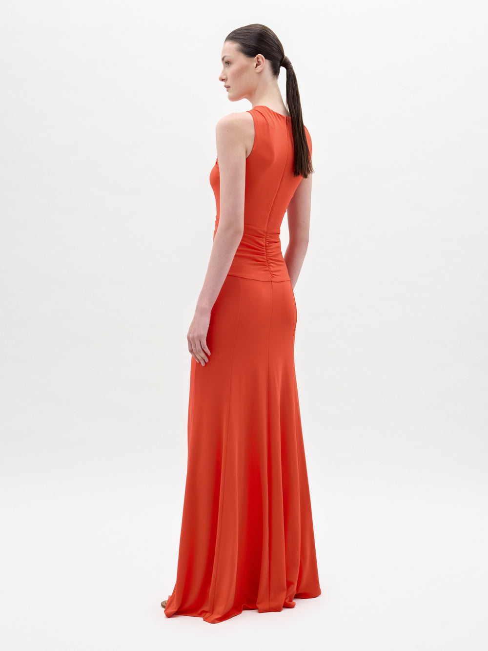 A woman stands against a plain white background wearing the Naga Dress, a long, sleeveless red maxi with a crossed halter neckline, front waist cutout, and an elegant column silhouette.