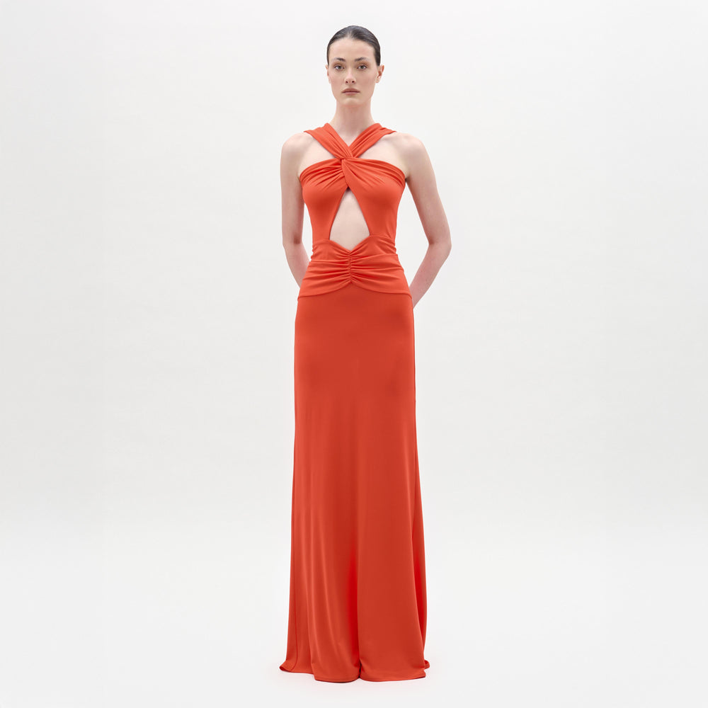 A woman stands against a plain white background wearing the Naga Dress, a long, sleeveless red maxi with a crossed halter neckline, front waist cutout, and an elegant column silhouette.