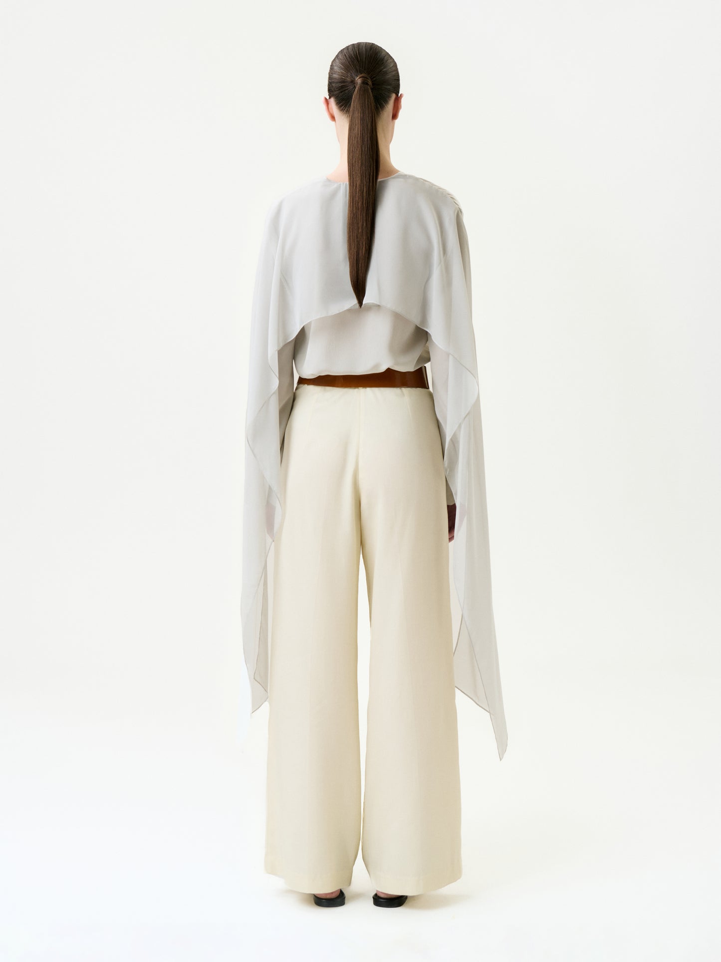 A person stands facing away in a flowy light grey Italian-fabric top with long draped sides, paired with the wide-legged Emine Pant in cream. Their hair is styled in a sleek low ponytail against a plain white background.