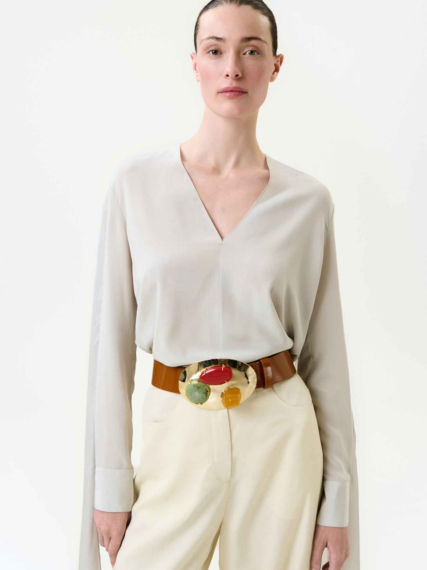 A woman with slicked-back hair wears the Vanna Blouse—a light beige, 100% silk Italian V-neck—paired with cream pants and a wide tan belt with a bold abstract buckle, standing against a plain white background.