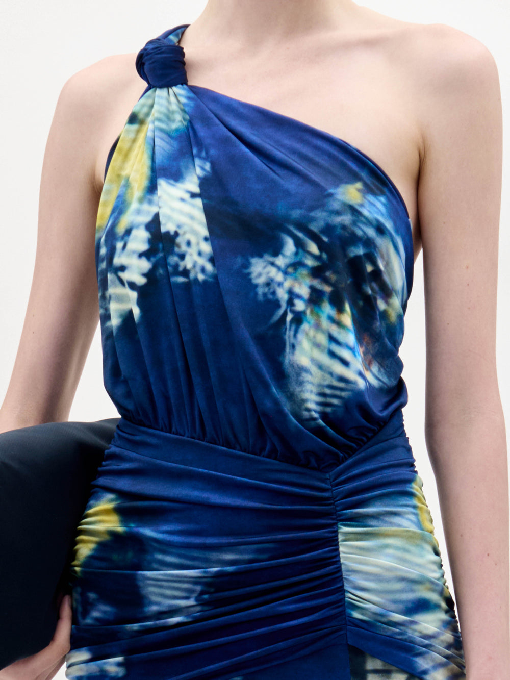 A woman stands against a plain white background wearing the Antara Dress, a one-shoulder, floor-length blue gown with yellow and white patterns, holding a black clutch.