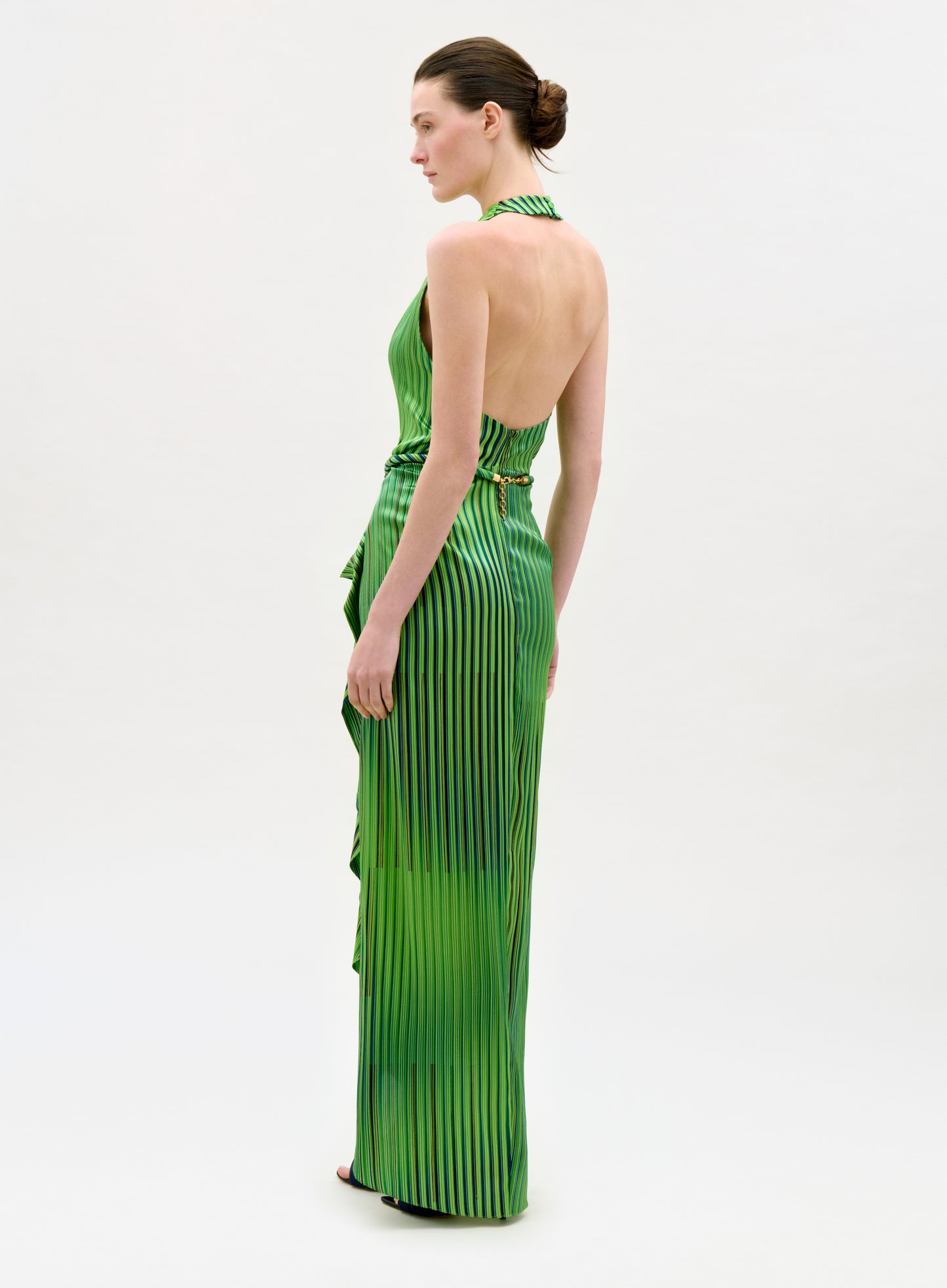 A woman in the Gypsy Dress, a sleeveless, backless green midi with vertical black stripes and an A-line silhouette, stands sideways against a plain light background. Her hair is tied back in a neat bun.