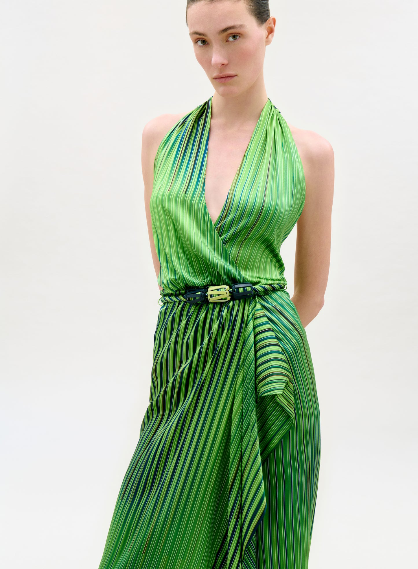 A woman stands against a plain background, wearing the Gypsy Dress—a sleeveless, green halter-neck midi dress with diagonal stripes, an A-line silhouette, a gathered skirt, and a cinched waist with a dark belt.