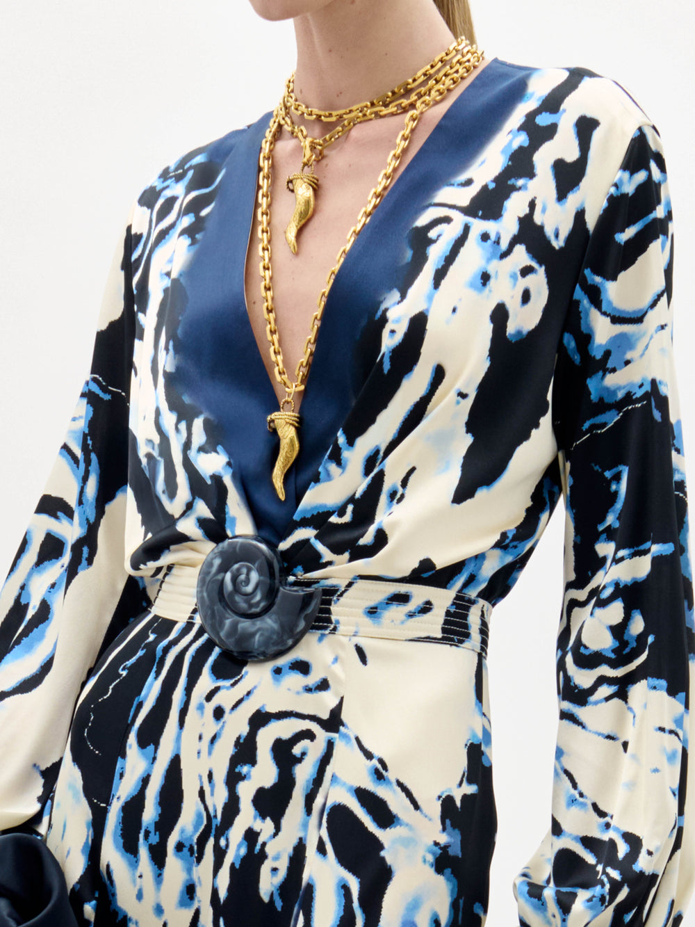 A woman wearing the Remy Dress—a long, patterned dress in blue and cream—holds a black fringed bag while standing against a plain white background.