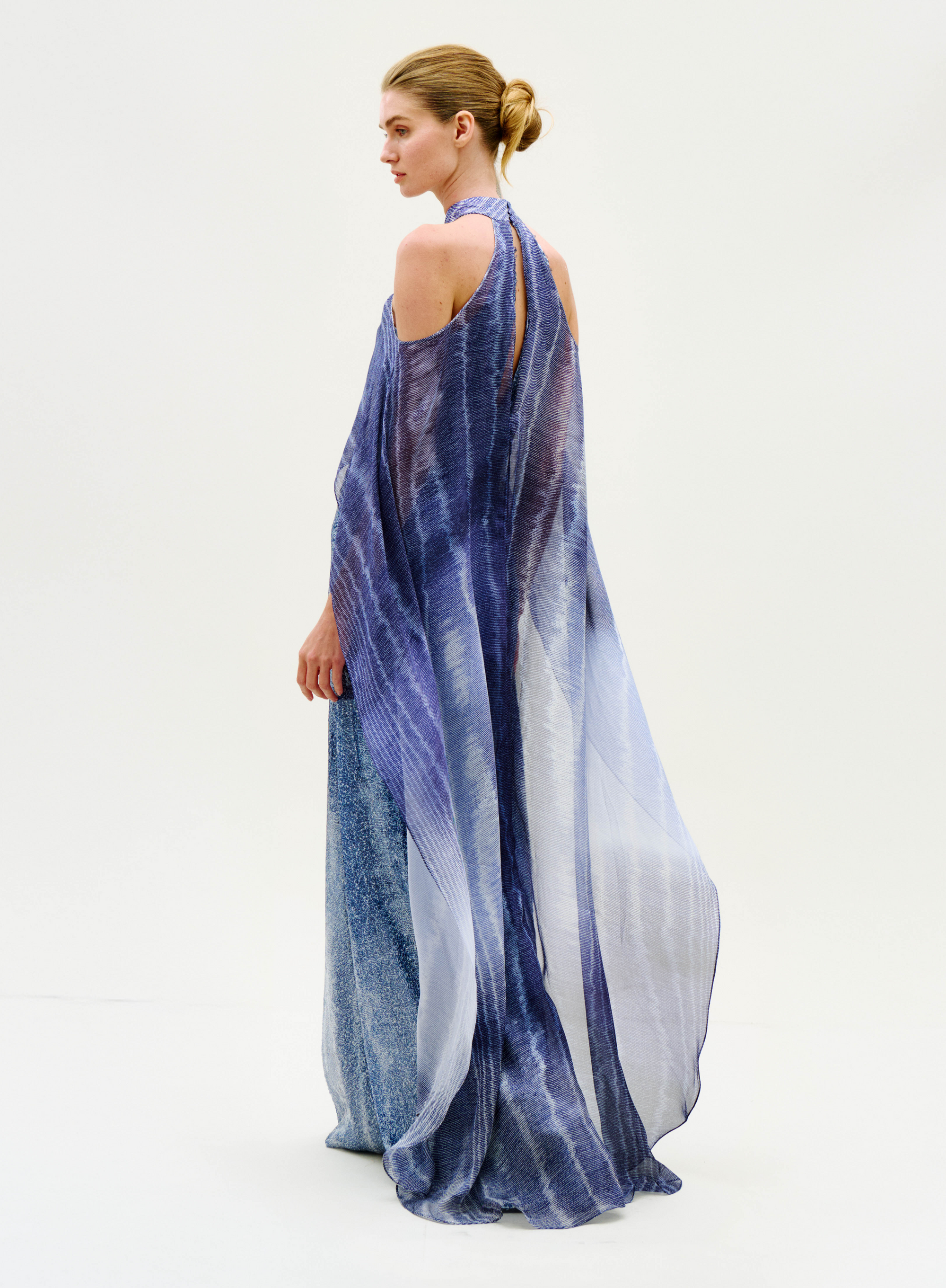 Wearing the Serenela Dress, a person with hair in a low bun stands against a plain, light background, looking to the side. The flowing, sheer dress features open shoulders and comes in a blue-purple hue.