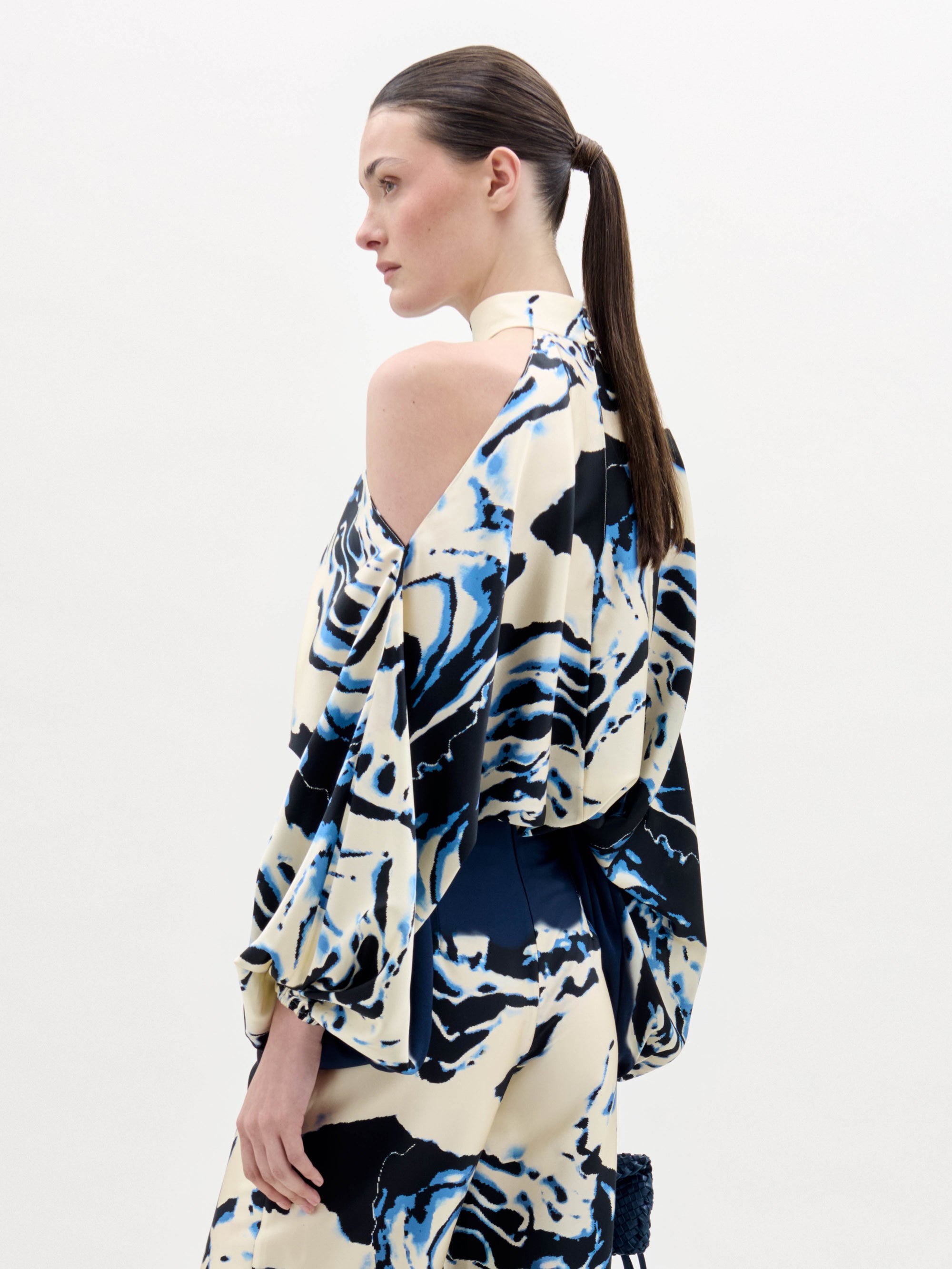 A woman stands against a plain background wearing the Safira Blouse, a high-neck, cold-shoulder top with matching wide-leg pants in a blue, black, and white marble pattern.