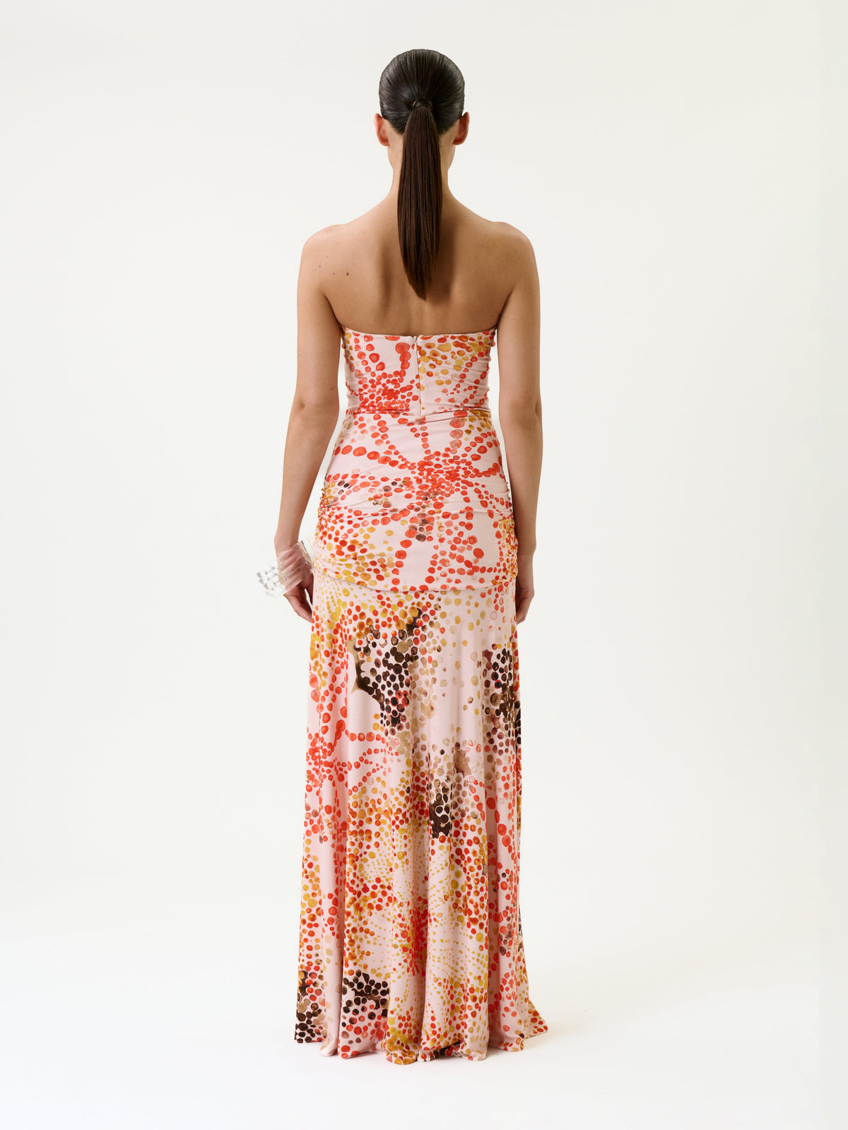 A woman with a ponytail stands facing away, wearing the Shani Dress—a strapless, floor-length gown featuring a colorful abstract dot pattern in red, pink, yellow, and brown tones. The background is plain white.