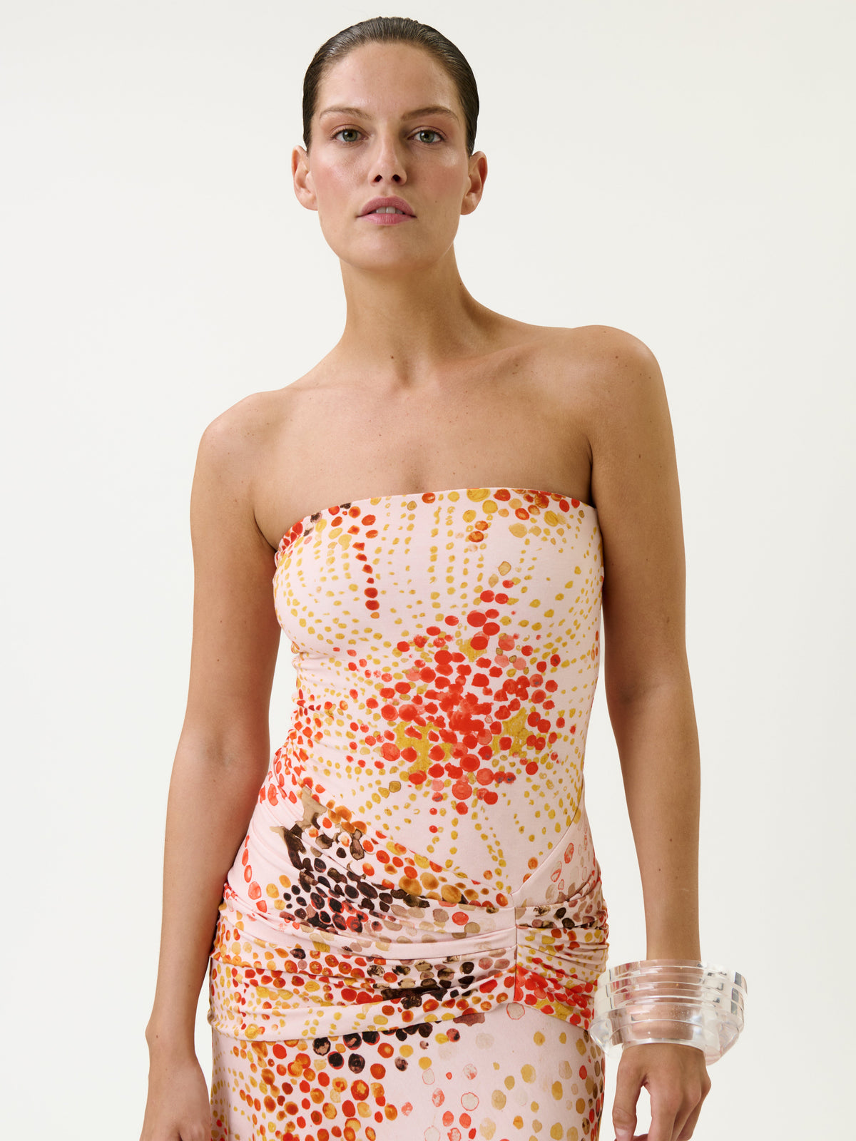 A woman with slicked-back hair wears the Shani Dress, a colorful, strapless piece featuring red, orange, and yellow dot patterns. She stands against a plain white background and has a clear, chunky bracelet on her right wrist.