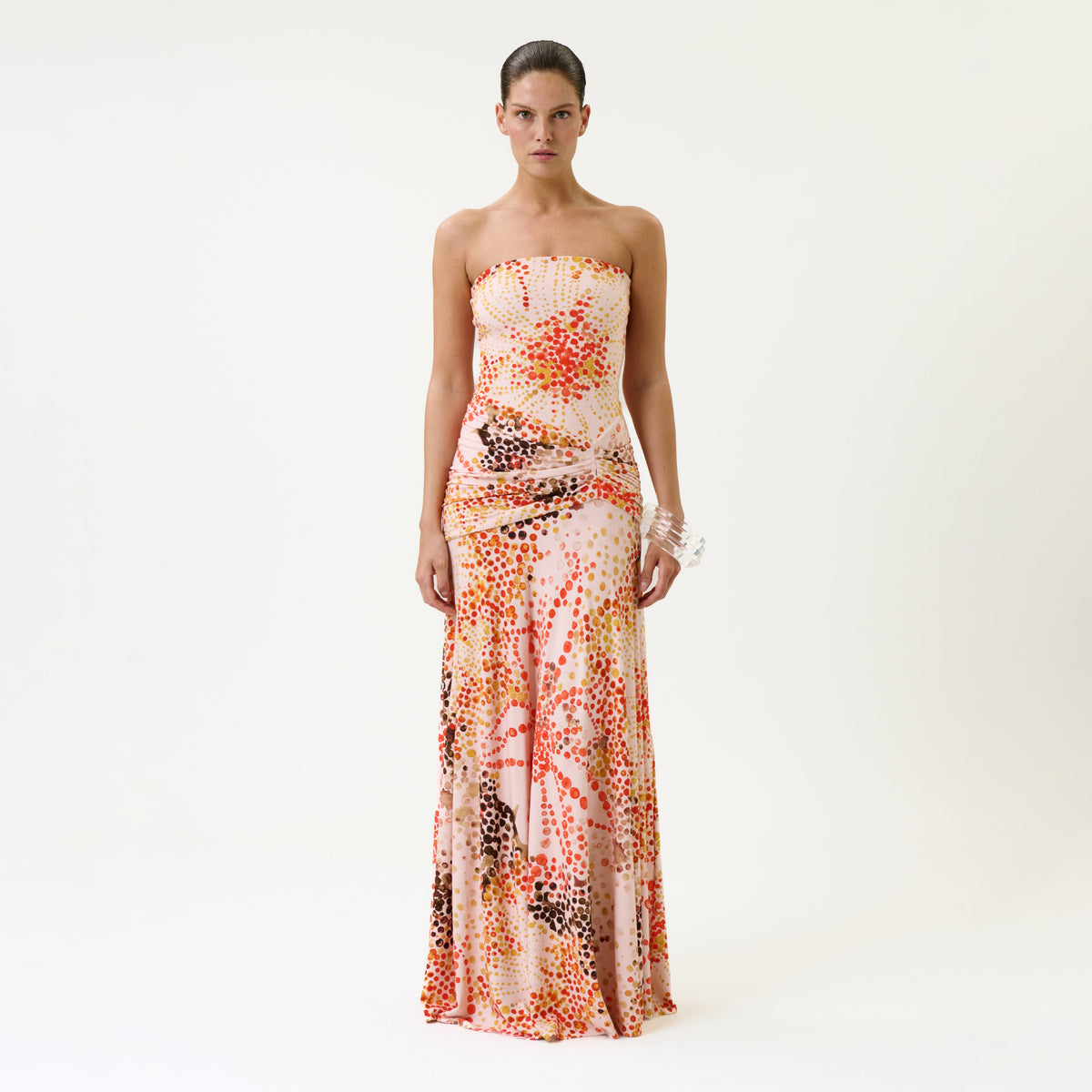 A woman stands against a plain white backdrop wearing the Shani Dress, a strapless, floor-length gown featuring a colorful abstract dot pattern in shades of orange, pink, and brown on a white background.