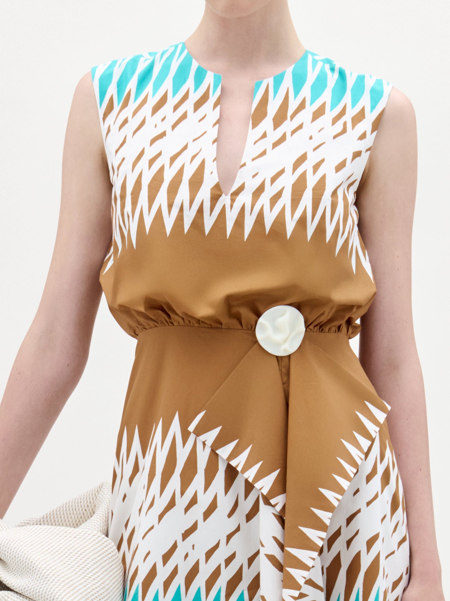 A woman stands against a white background wearing the Kendall Dress, a sleeveless turquoise, brown, and white fit-and-flare midi made from Italian fabric, paired with a large white bag and brown heeled sandals.