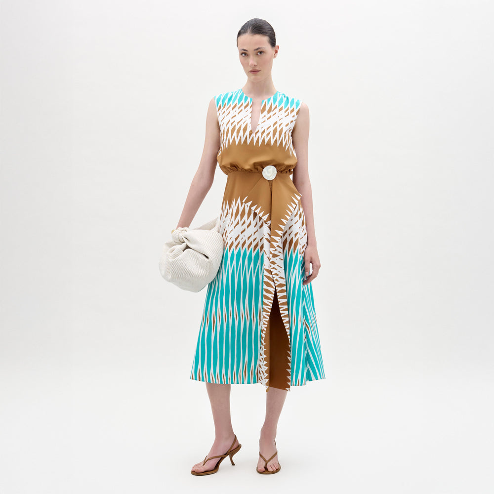 A woman stands against a white background wearing the Kendall Dress, a sleeveless turquoise, brown, and white fit-and-flare midi made from Italian fabric, paired with a large white bag and brown heeled sandals.