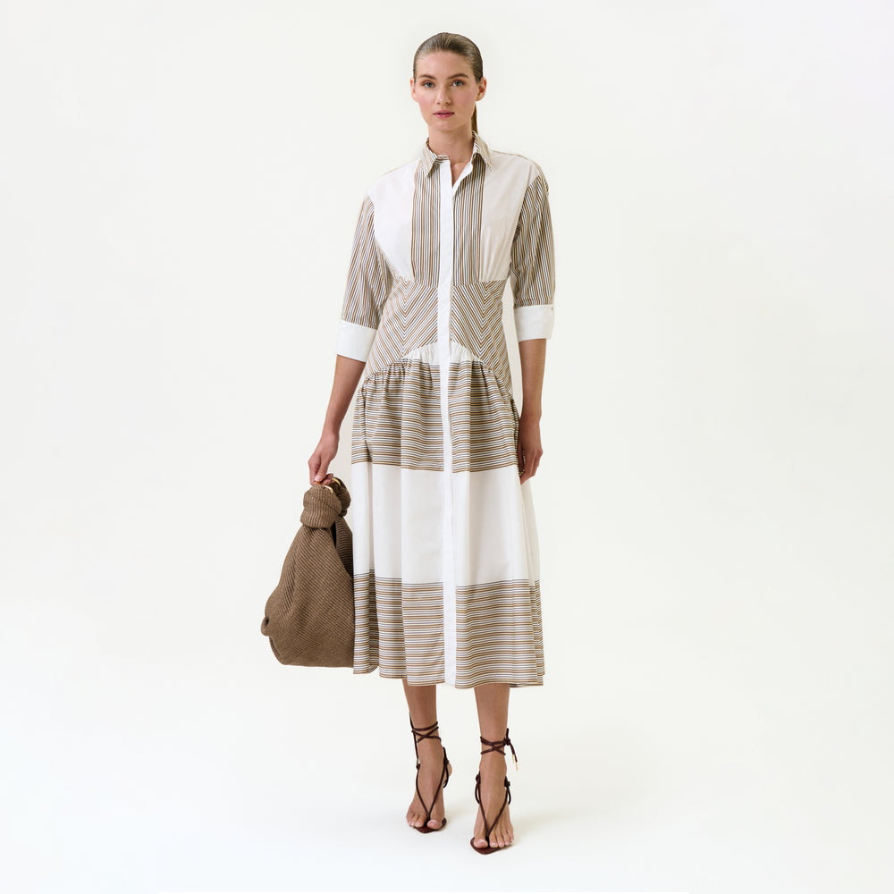 A woman stands against a white background wearing the Indy Dress—a long, collared midi dress with beige and white stripes in 100% Italian cotton. She carries a large brown textured bag and pairs the look with black strappy sandals.