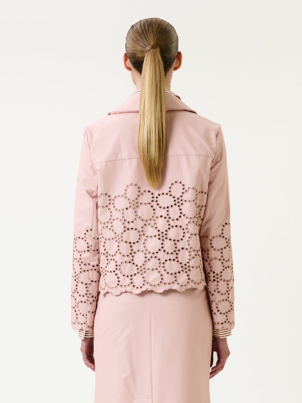 A woman with long, straight blonde hair in a ponytail is seen from the back against a plain white background, wearing the Belen Jacket—a light pink piece with eyelet detailing—paired with a matching pink skirt.