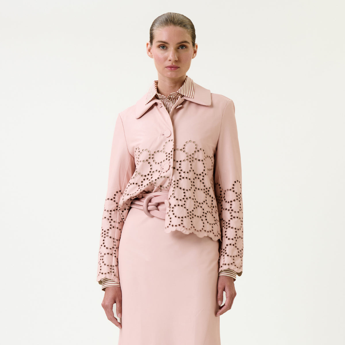 A woman stands against a plain background, wearing the Belen Jacket—a light pink piece featuring circular cut-out patterns on the sleeves and lower half—paired with a matching skirt over a striped collared shirt. Her hair is slicked back.