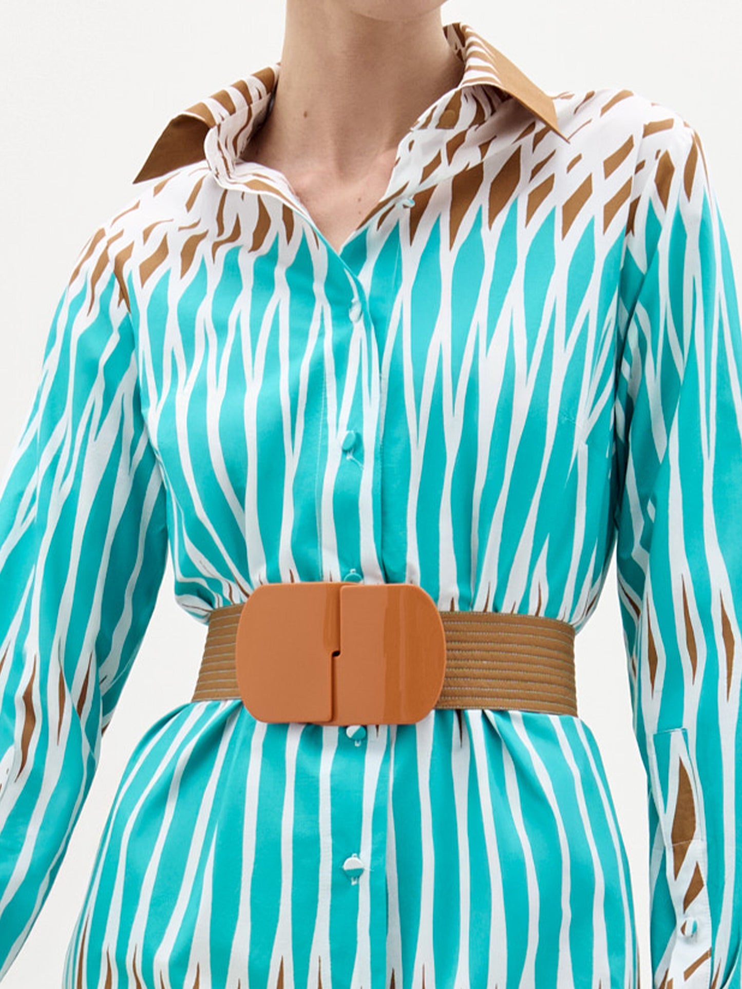 A woman stands against a plain background wearing the Ronzona Dress—a blue and brown patterned Italian midi dress—styled with a brown detachable belt, brown sandals, and holding a large white clutch.