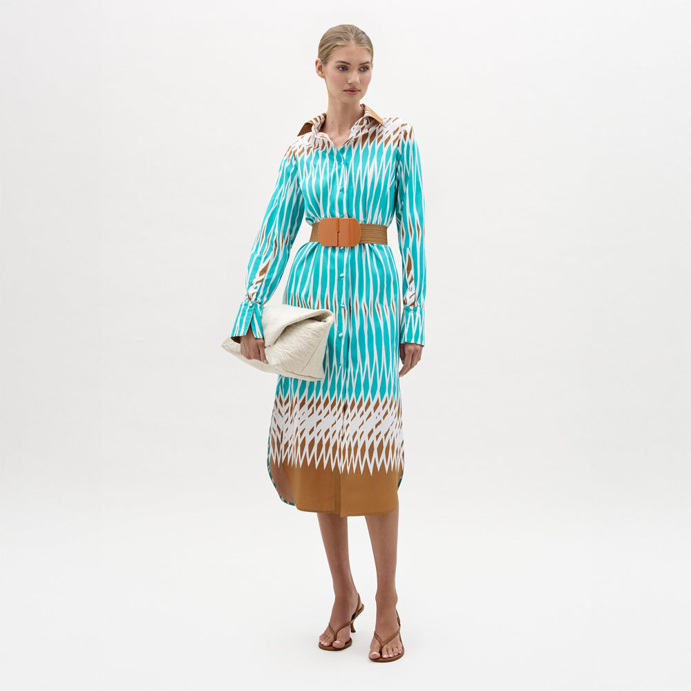 A woman stands against a plain background wearing the Ronzona Dress—a blue and brown patterned Italian midi dress—styled with a brown detachable belt, brown sandals, and holding a large white clutch.
