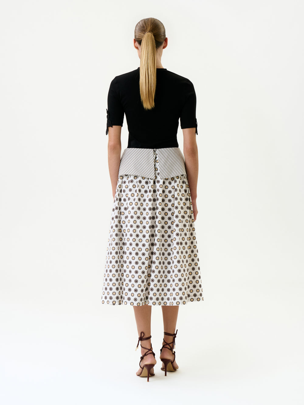A woman with a low ponytail faces away, wearing a fitted black top and the Maxima Skirt—an elegant, high-waisted, mid-length white cotton skirt with geometric patterns. She pairs it with brown strappy heeled sandals against a plain white background.